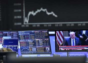 Donald Trump with computer screens displaying fluctuating stock graphs and trade data, representing the market impact of the 2025 U.S. tariff policies.